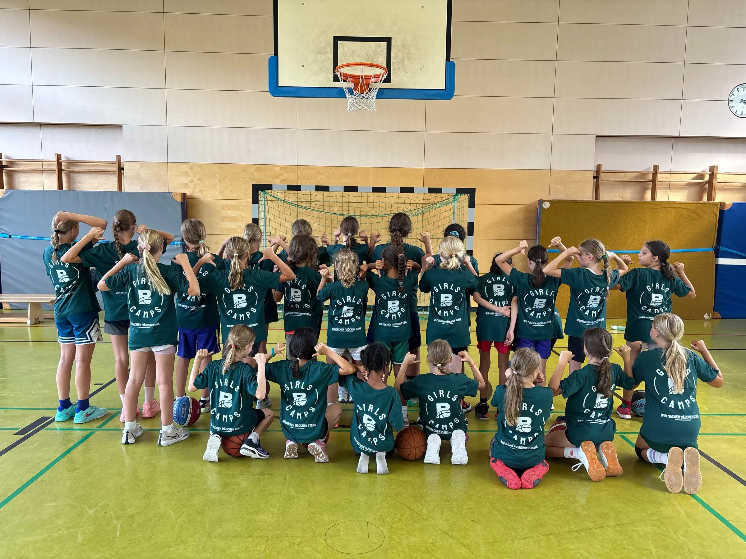 Rund zwanzig Mädchen in grünen Shirts mit dem Aufdruck „Girls Camps“ stehen und knien von hinten gesehen in der Halle und zeigen mit den Daumen auf den Schriftzug auf ihrem Rücken.