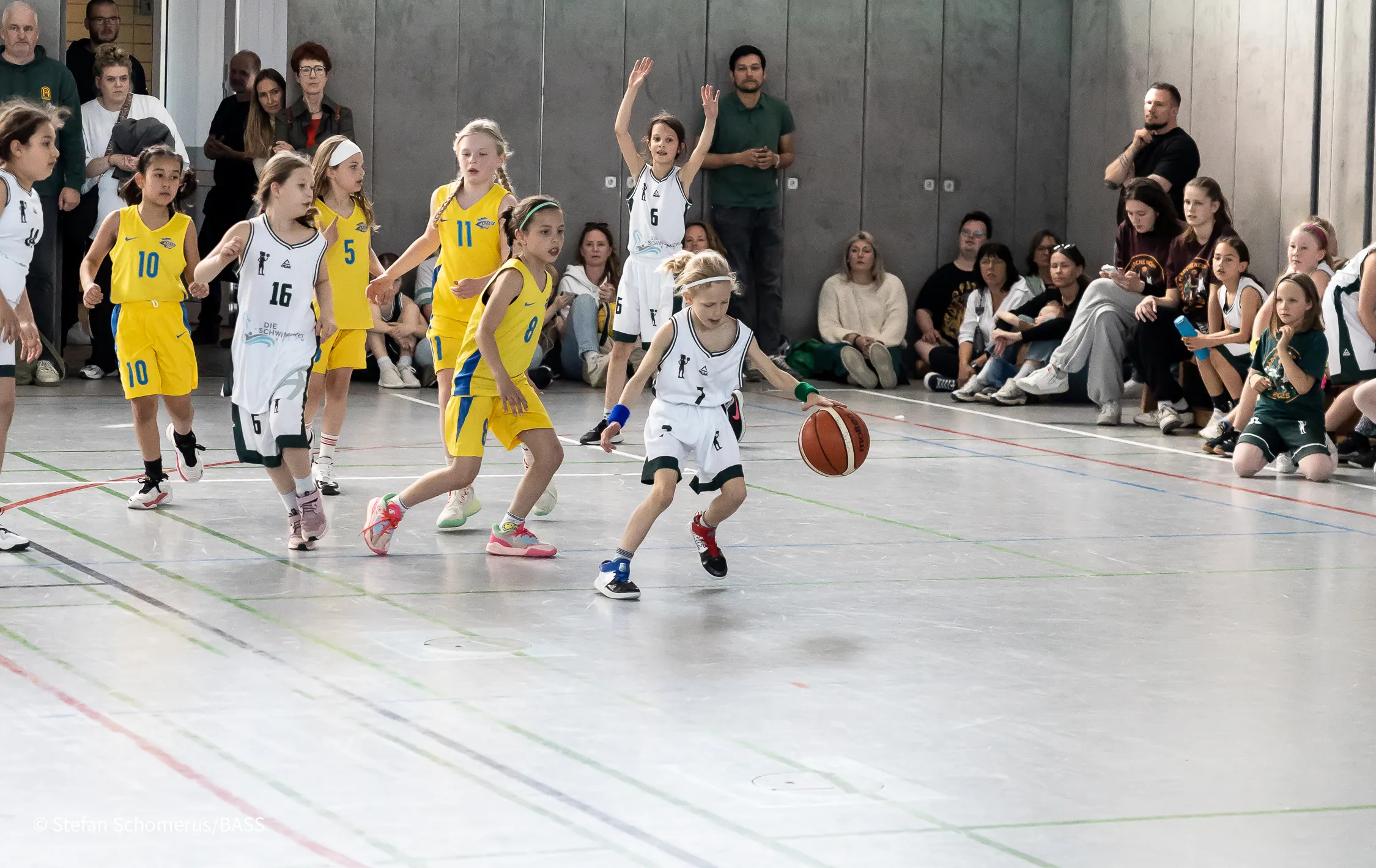 Spielszene beim BASS Mädchencup: eine junge Spielerin in Weiß dribbelt, Gegenspielerinnen in Gelb laufen mit, am Hallenrand sitzen und stehen dicht gedrängt Zuschauer*innen.